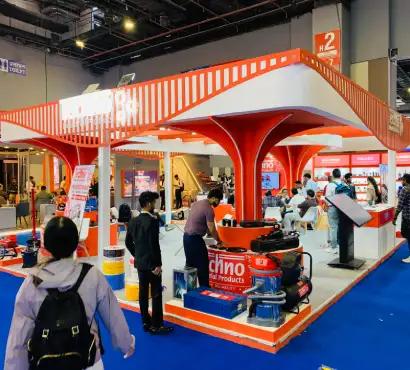 Wide-angle view of an industrial exhibition booth with orange and white branding, product displays and visitor areas